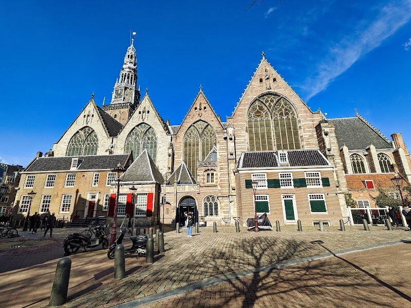Oude Kerk Amsterdam - Oude Kerk, Amsterdam’s oldest building, stands at the heart of the city’s vibrant Red Light District, offering families and curious travelers a fascinating blend of history and culture. Founded in the early 13th century as a modest wooden chapel, it was rebuilt and expanded over centuries into the grand Gothic and Dutch Renaissance structure you see today. Step inside and you’ll be greeted by the largest medieval wooden vault in Europe, a breathtaking ceiling crafted from Estonian oak dating back to 1390, renowned for its exceptional acoustics. The church’s floor—unlike any other—is paved entirely with gravestones, marking the resting places of 60,000 Amsterdam citizens, including notable figures such as Rembrandt’s beloved wife Saskia van Uylenburgh and composer Jan Pieterszoon Sweelinck. Kids and adults alike will be intrigued by the contrast between the tranquil interior and the lively neighborhood outside, as well as the stories embedded in the church’s walls. Visitors can marvel at the stunning stained glass, explore the sculpted choir stalls, and even attend organ concerts on the historic 17th-century grand organ. Whether you’re seeking a quiet moment, a dose of Amsterdam’s history, or a unique family outing, Oude Kerk promises a memorable experience for all ages.