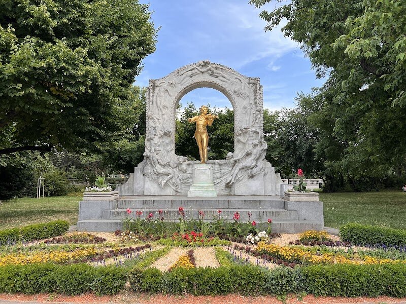 Johann Strauss monument - Standing majestically in Vienna's Stadtpark is one of the world's most iconic and photographed monuments—the gleaming golden statue of Johann Strauss II, the legendary "King of the Waltz." Sculptor Edmund Hellmer created this masterpiece, which was unveiled on June 21, 1921, more than two decades after Strauss's death. The statue captures the composer in his element: violin in hand, poised elegantly before his orchestra, framed by a stunning marble arch adorned with dancing figures. What makes this monument truly special is its radiant presence—the bronze figure is gold-plated, catching the light beautifully whether you visit by day or experience it illuminated after dark. This is where Strauss's genius comes alive, the man who gave Vienna the "Blue Danube," Austria's beloved anthem. The monument sits just behind the historic Kursalon concert hall, where Strauss performed his first concert in 1868. Today, visitors can stroll through the tree-lined park, soak in the musical heritage, and even enjoy waltz concerts at the nearby Kursalon, bringing Strauss's timeless compositions to life in the very spaces that inspired him.